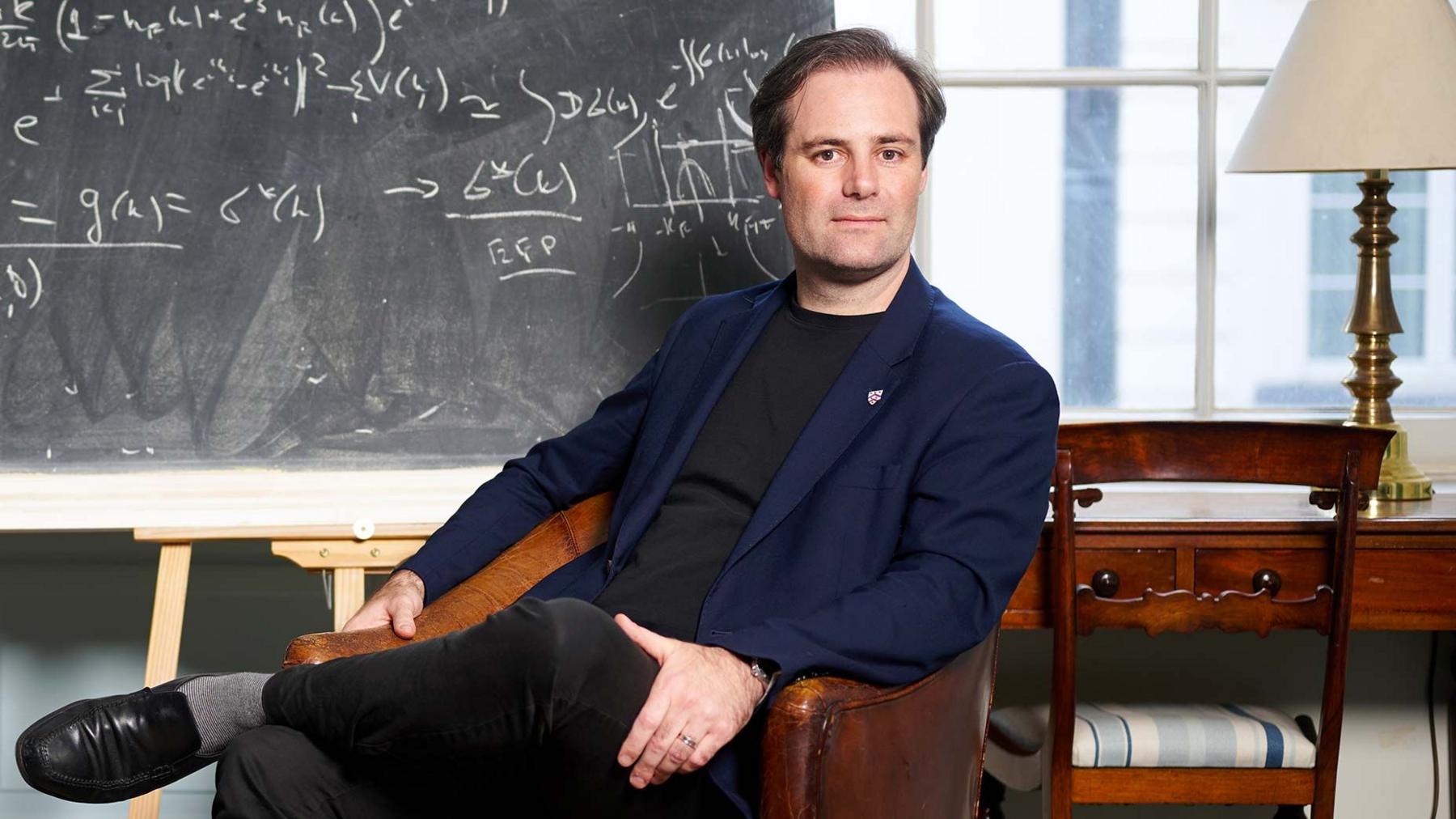 Ben Delo sitting ona leather armchair, a blackboard with mathematical equations writen in chalk in  behind him 