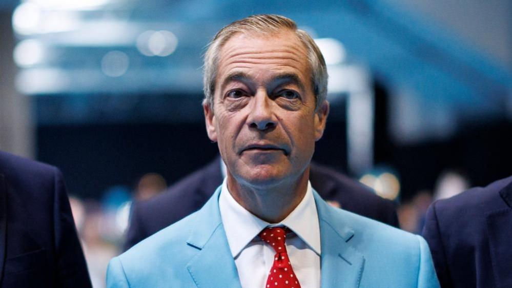 Reform UK leader Nigel Farage is wear a turquoise-coloured suit jacket, white shirt and a red tie with white dots.