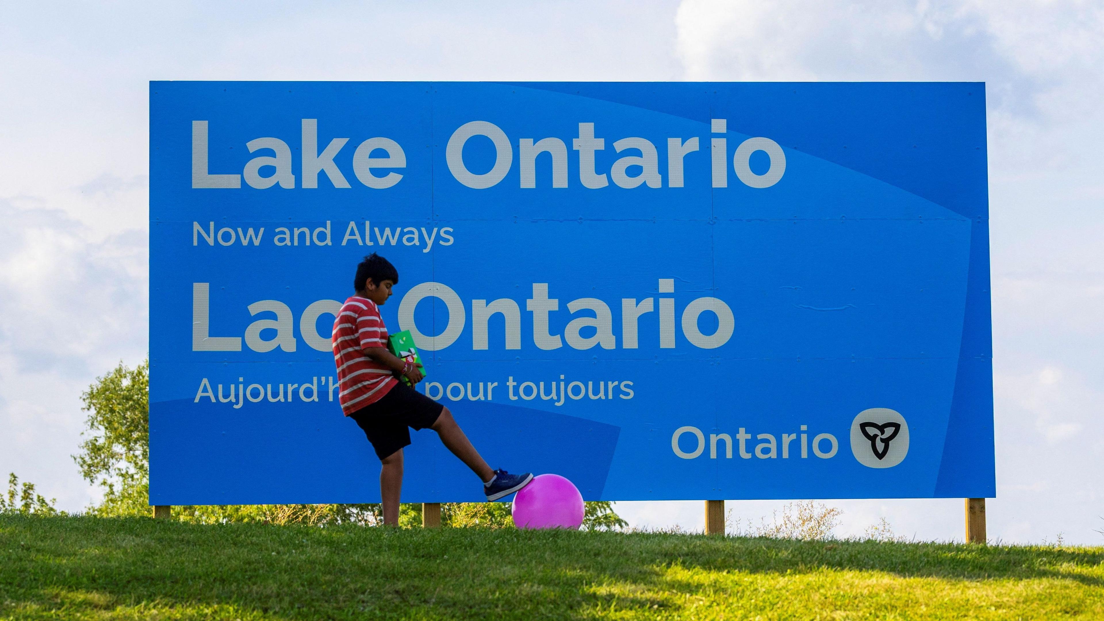 A sign reads "Lake Ontario now and always" with a boy playing with a ball nearby. 