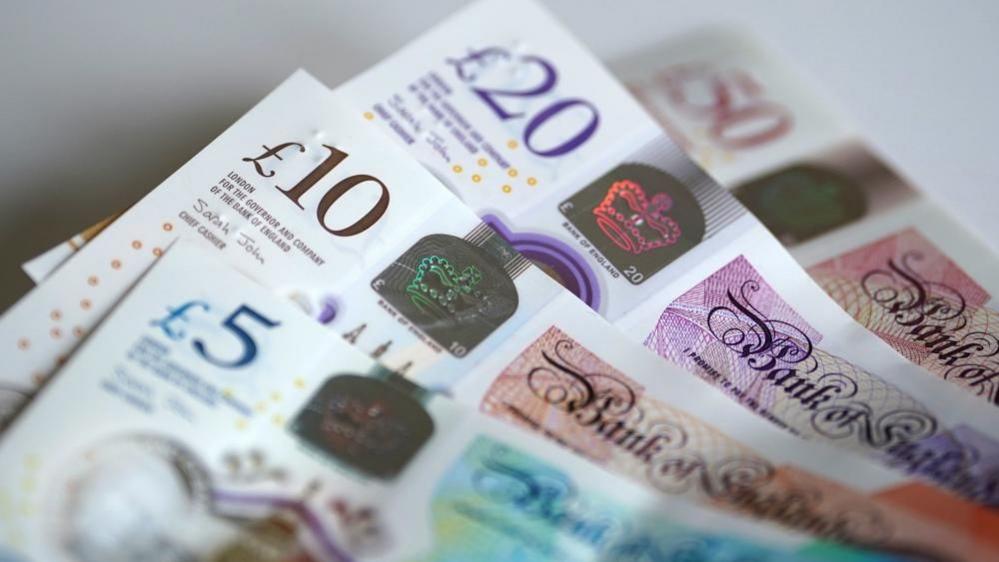 Four banknotes are in a pile on a table. Each banknote is partially obscured. There is a £50 note in the background which has a £20 on top of it. A £10 is on top of the £20, and a £5 is on top of the £10.