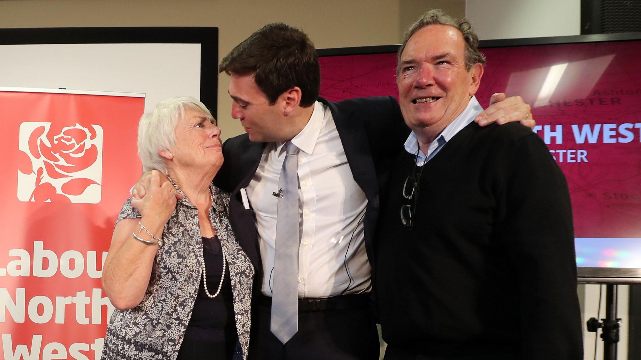 Andy Burnham hugs his parents, Eileen and Roy, after he was selected as the Labour candidate to become the mayor of Greater Manchester in September 2026. They are standing in front of a red banner reading "Labour North West".
