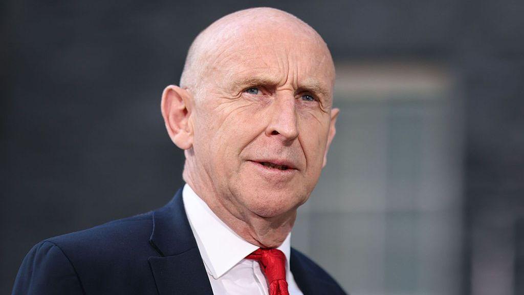 John Healey stands looking into the middle distance wearing a red tie, white shirt and navy blue suit. There is a blurred window behind him.