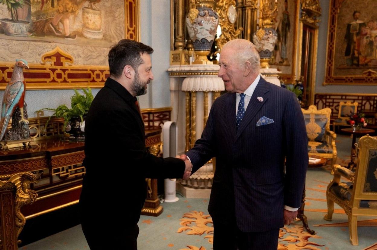 Volodymyr Zelensky and King Charles smiling at each other while shaking hands inside a grand room in Buckingham Palace.