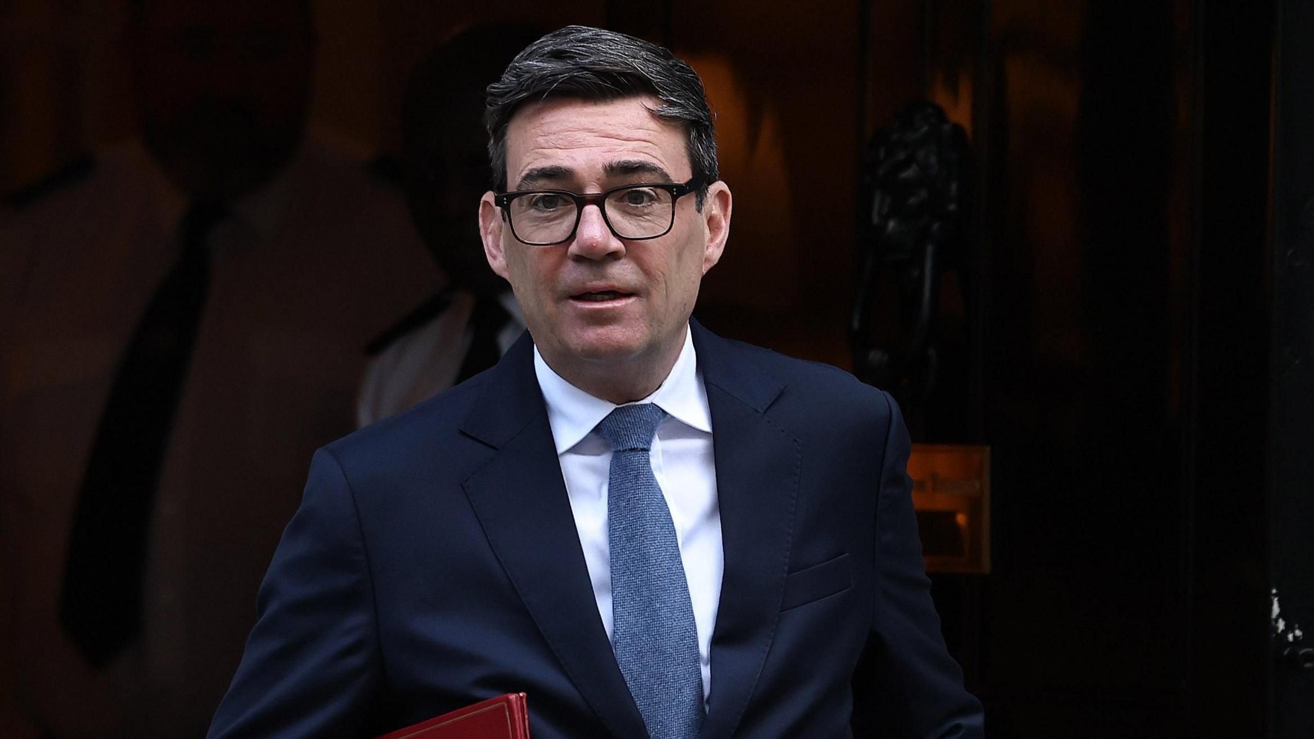 Andy Burnham leaving Downing Street. He is wearing a dark blue suit, blue tie and glasses. He is carrying a red folder. 