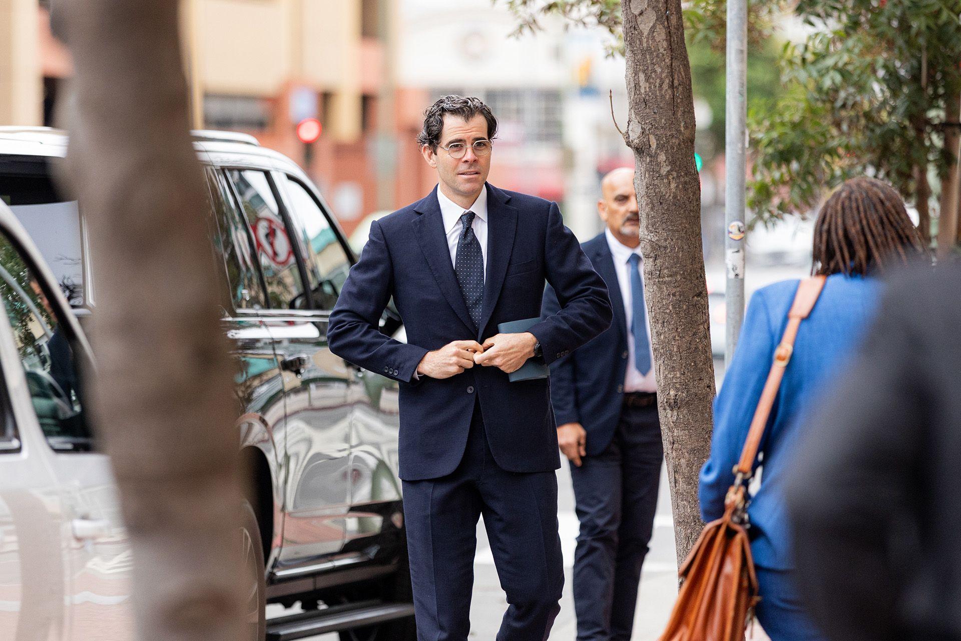 Adam Mosseri, wearing a navy suit and spotted tie, walks in front of one of his entourage in front of cars on a street while attending court in California in August.