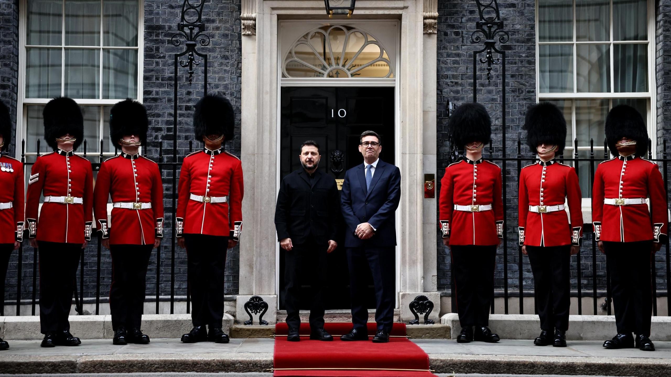 Ukrainian President Volodymyr Zelensky and UK Prime Minister Andy Burnham standing in front of No 10 Downing Street. They are standing on a red carpet. On either side of them are soldiers from the Number 12 Company of the Irish Guards, wearing scarlet tunics and bearskin hats.
