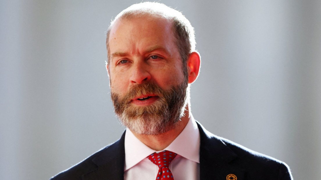 Head and shoulders shot of Reynolds, wearing a black jacket over a white shirt and red tie. The backdrop is out of focus and pale but sunny.