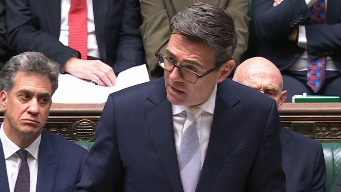 Prime Minister Andy Burnham making a statement on international affairs in the House of Commons. He wears a navy suit with a white shirt and a pastel tie with broad grey, lilac and cream stripes. HIs black hair is swept sideways and he wears dark brown spectacles.