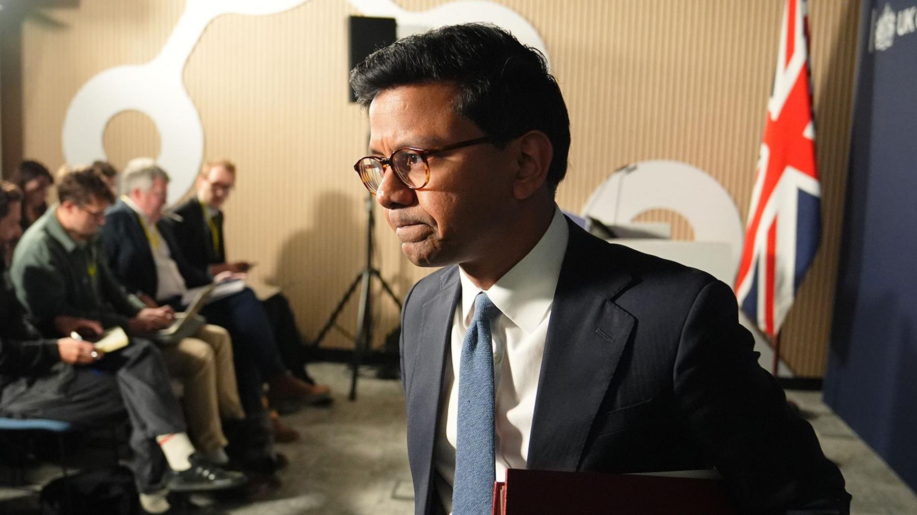 Kanishka Narayan walking away from podium after speaking  in a suit with a blue tie against a union jack flag backdrop and blue background and memebers of te press behind him