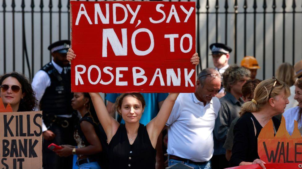 A woman holding a red sign reading "Andy, say no to Rosebank" at a protest 