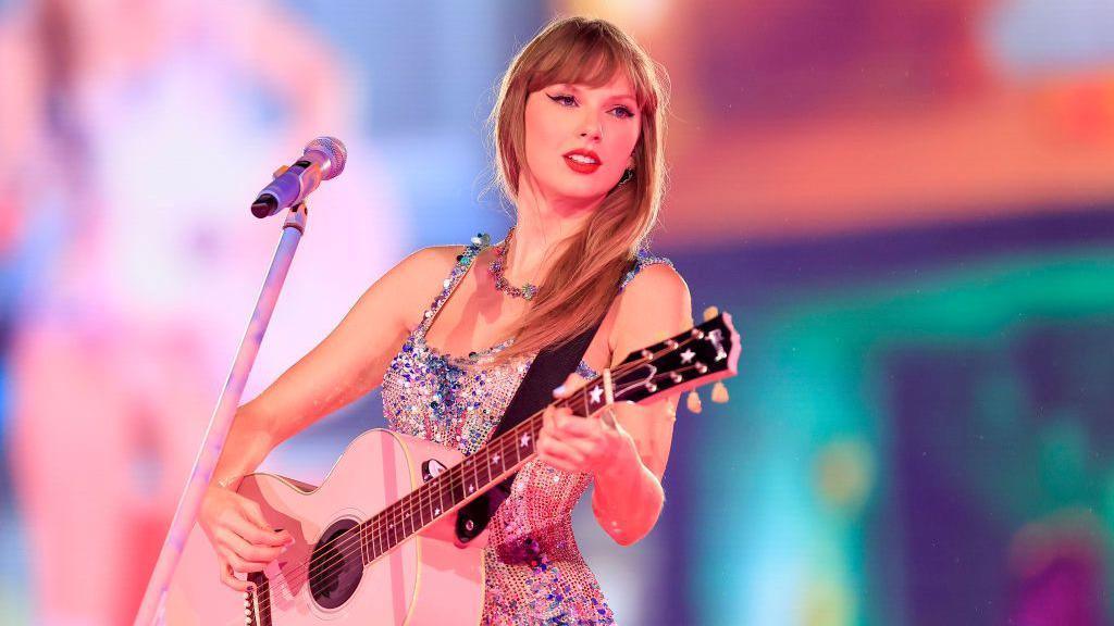 Taylor Swift on stage playing a pink guitar in a sparkly top