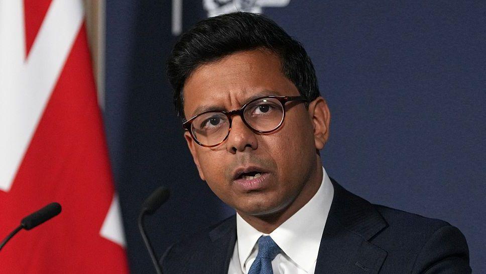 Kanishka Narayan speaking in a suit with a blue tie against a union jack flag backdrop and blue background